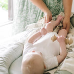Baby lying in a sage green baby nest lounger with a person in a green dress interacting nearby